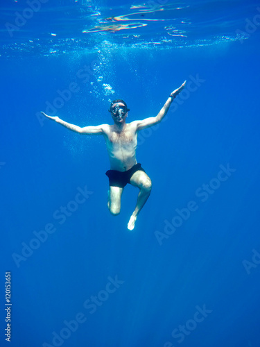 Portrait of a young causasian man meditating in the lotus position underwater. Submerged under water free diving, crossed legs, wearing a mask and blowing bubbles.