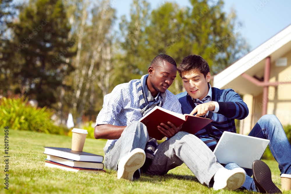 Two young students sitting on grass in campus and looking through a textbook Two young students sitting on grass in campus and looking through a textbook