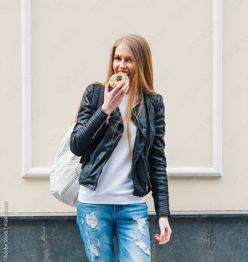 Beautiful girl bites a delicious donut walking on the streets of European city. Warm spring. Outdoor Beautiful girl bites a delicious donut walking on the streets of European city. Warm spring. Outdoor