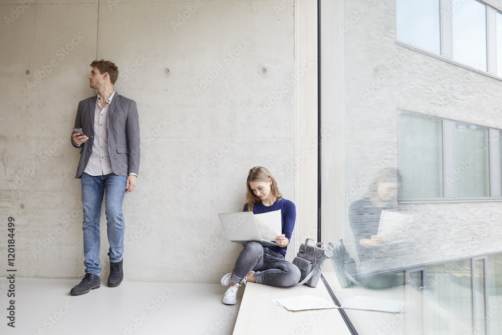 Young woman with papers and laptop and man with cell phone at concrete wall Young woman with papers and laptop and man with cell phone at concrete wall