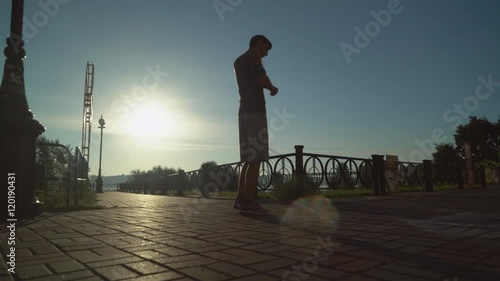 A young man wearing in shorts and T-shirt listening music with headphones and begins to run forward. Urban city view in sunrise. Jogger running in the morning at the summer season. On the background photo