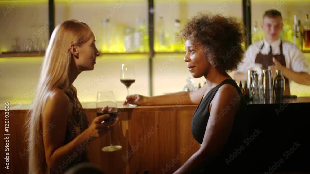 Two girls chatting and drinking wine at the luxury bar. At background barman makes cocktails