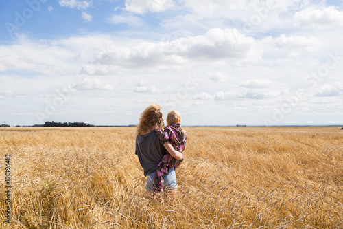 A teenage girl holding her toddler sister standing in a ripe crop of golden wheat under cloudy countryside landscape
