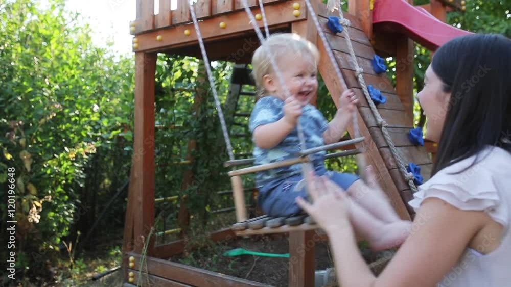 One years-old baby boy with blue eyes play on the swing outdoor on a sunny day. Mum helps the little son swinging on the swing. slowmotion One years-old baby boy with blue eyes play on the swing outdoor on a sunny day. Mum helps the little son swinging on the swing. slowmotion
