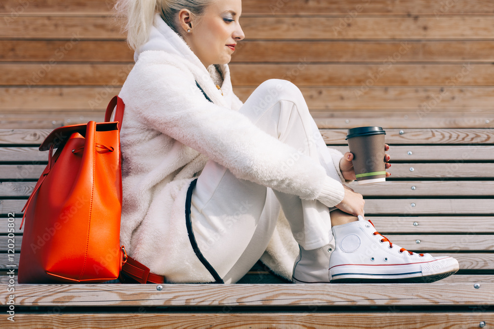 Φωτογραφία Girl sitting on the bench with a big red super fashionable backpack in the white sports suit and sneakers with cup of coffee on a warm summer evening