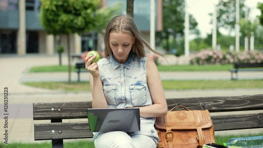 Pretty student eating apple and studying on laptop while sitting on the bench