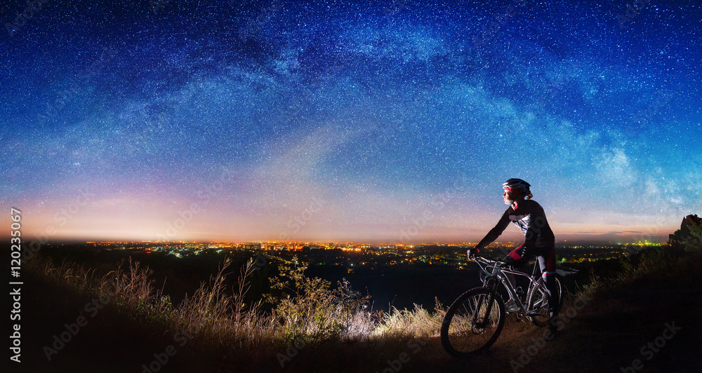 Obraz premium Cross country biker on top of the hill observing beautiful panoramic view of night sky and city lights