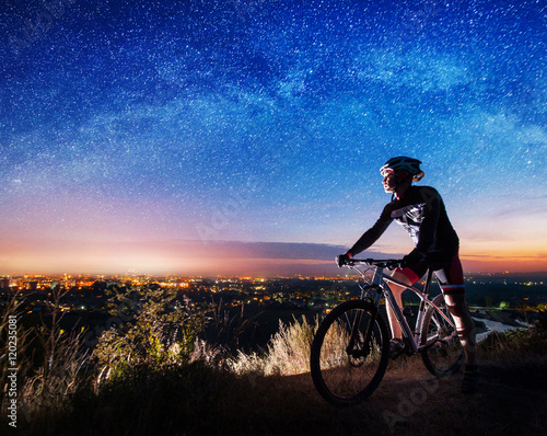 Young athlete biker with mountain bike on top of the hill observing the evening city view under starry sky