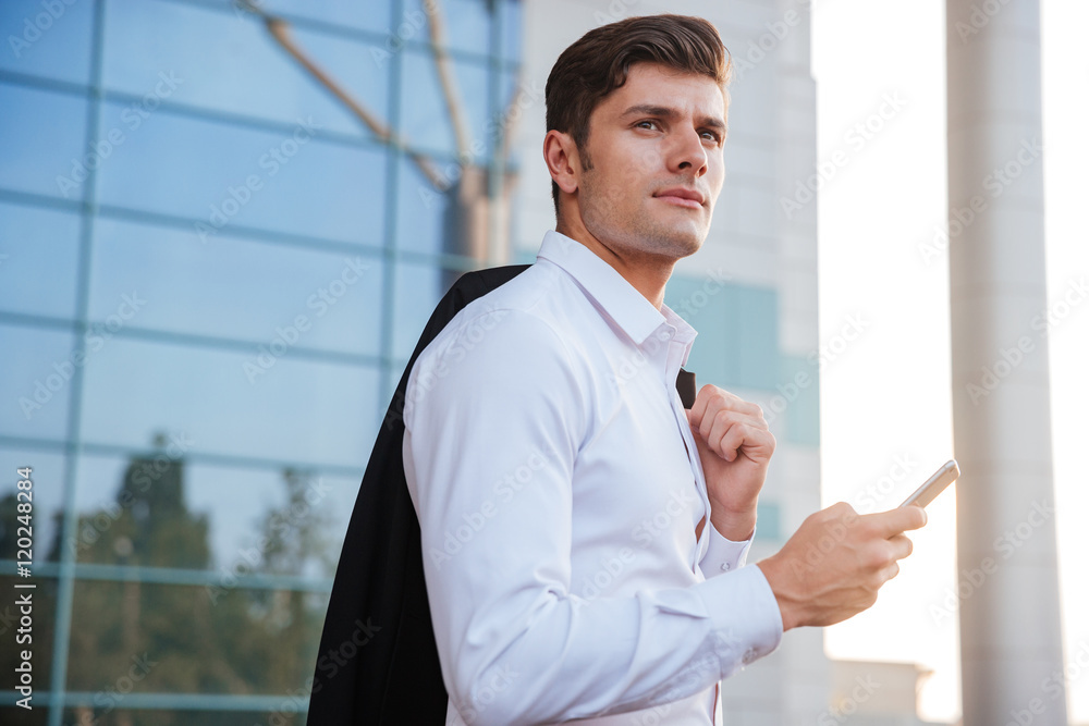 Pensive young businessman holding mobile phone with jacket over shoulder Pensive young businessman holding mobile phone with jacket over shoulder