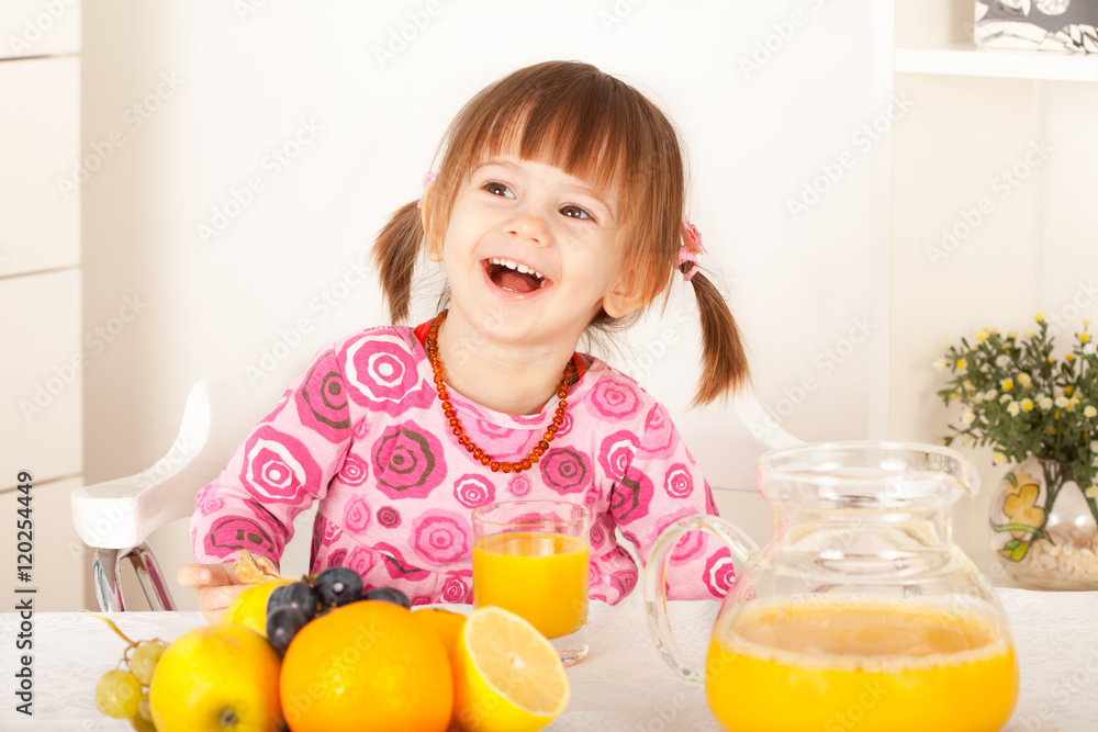 Cute little girl with pigtails sitting and laughing. Fruit and orange juice on the table. Cute little girl with pigtails sitting and laughing. Fruit and orange juice on the table.