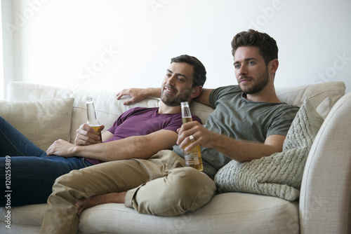 Gay Male Couple On Sofa At Home Watching TV Drinking Beer