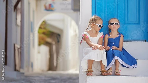 Two girls in blue dresses having fun outdoors. Kids at street of typical greek traditional village with white walls and colorful doors on Mykonos Island, in Greece