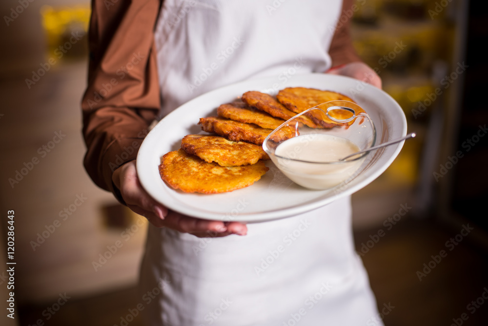Potato pancakes served on white plate with sour creamon a white plate in hand of woman with white apron. Close-up Potato pancakes served on white plate with sour creamon a white plate in hand of woman with white apron. Close-up