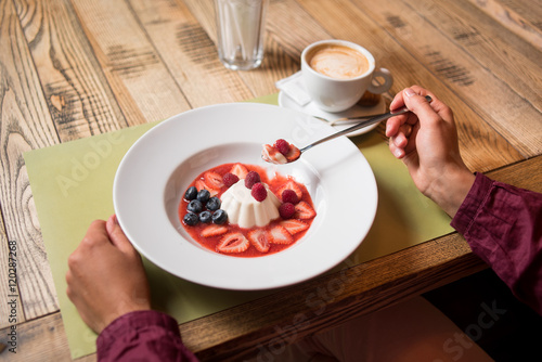 Woman eating white milky creamy pudding with jam and berries blueberries, strawberry, raspberry on table in cafe or restaurant with cup of coffee. Close-up