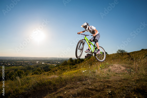 Man making extreme jump on a mountain bike on the hill against blue sky  sun and greenery into the distance. Cyclist is wearing white sportswear helmet and glasses.