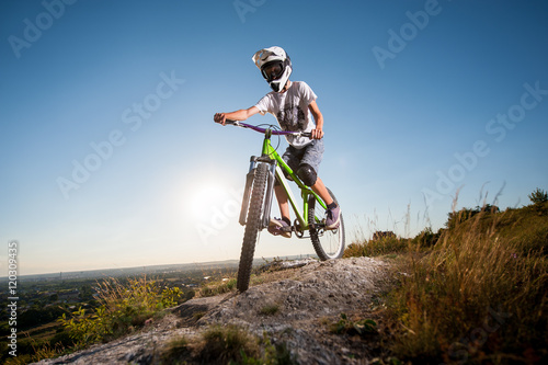 Fototapeta Naklejka Na Ścianę i Meble -  Male bicyclist in helmet and glasses riding on the mountain bike at the hill under blue sky in the mountains. Downhill cycling. Wide angle view