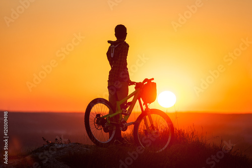 Silhouette of the guy with the bike at the top of mountain on the sunset background. Cyclist looking to the evening bright sun