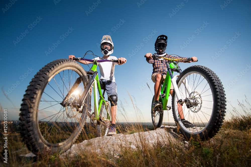 Two cyclists wearing in helmets and glasses standing with the mountain bikes on the top of the mountain under blue sky. Men looking into the camera. Bottom view Two cyclists wearing in helmets and glasses standing with the mountain bikes on the top of the mountain under blue sky. Men looking into the camera. Bottom view