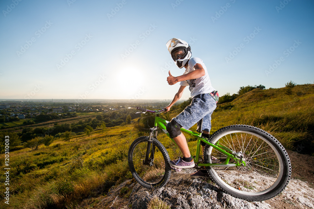 Bicyclist in helmet and glasses on mountain bike stands on the precipice of hill and showing thumb up gesture of good class under blue sky and sun. Wide angle view
