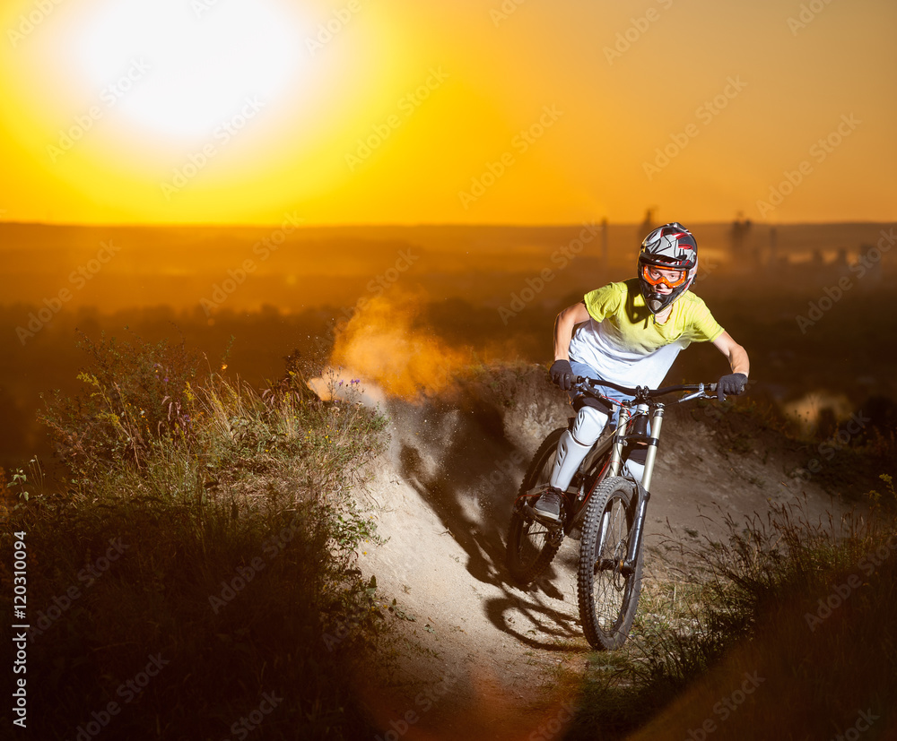 Naklejka premium Young rider in helmet and glasses is riding on the mountain bicycle on a dusty trail against evening sky with bright sun on the sunset. Blurred background