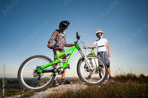 Two guys wearing in helmets and glasses standing with the mountain bikes on the hill under blue sky and looking into the camera. Bottom view