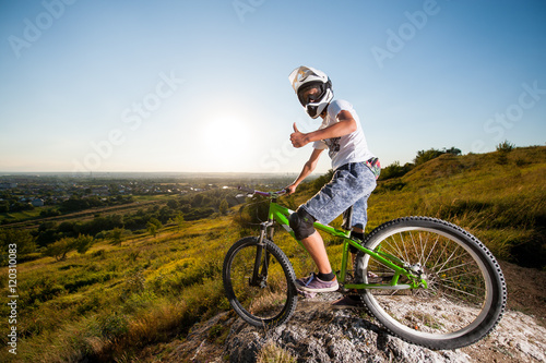 Bicyclist in helmet and glasses on mountain bike stands on the precipice of hill and showing thumb up gesture of good class under blue sky and sun. Wide angle view