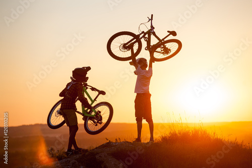 Two guys in helmets holding a mountain bikes in hands on the top of the mountain against evening sky with bright sun at the sunset