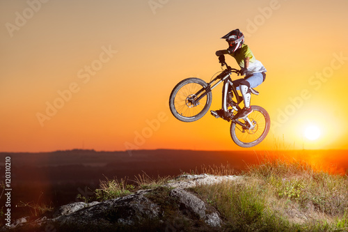 Fototapeta Naklejka Na Ścianę i Meble -  Sunset. Biker flying on a mountain bike on the precipice of hill against evening sky with bright sun. Cyclist is wearing sportswear helmet and glasses. Extreme sport