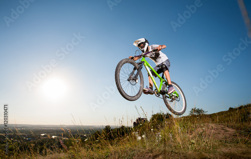 Athlete biker jumps on a mountain bike on the hill against blue sky into the distance in the evening. Cyclist is wearing white sportswear helmet and glasses.