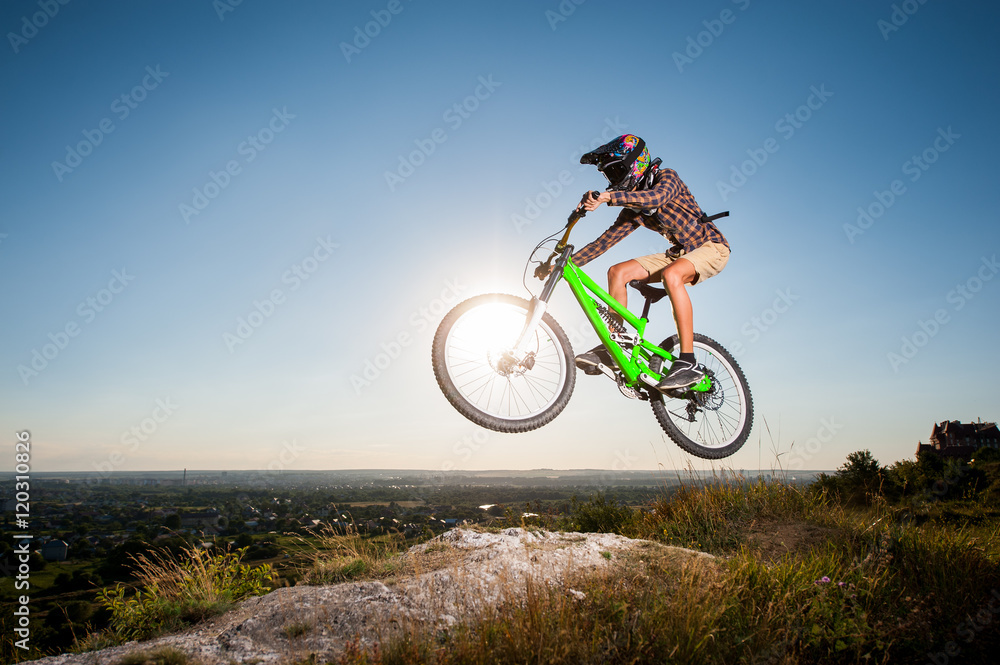 Bicyclist riding downhill and making extreme jump on a mountain bike into the distance from the slope in the mountains against blue sky with sun. Wide angle view