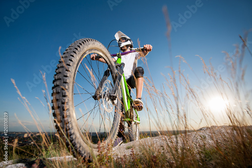 Cyclist in helmet and glasses stay on the bicycle at the hill under blue sky and sun and looking  into the distance. Focus on the bike. Bottom point view