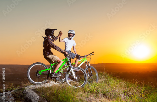 Guys in helmets and glasses stay on the bicycles at the precipice of hill and give high five for each other against evening sky with bright sun at the sunset. Blurred background
