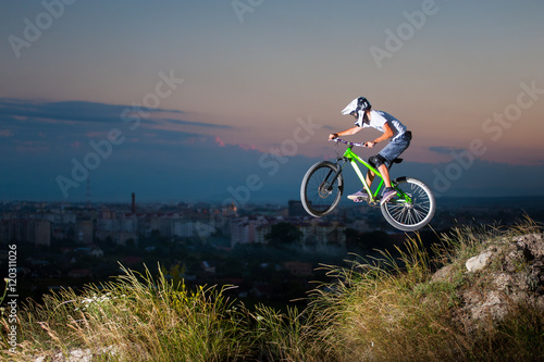 Rider in helmet and glasses flying on a mountain bike on the hill against evening sky and small town into the distance. Extreme downhill cycling.