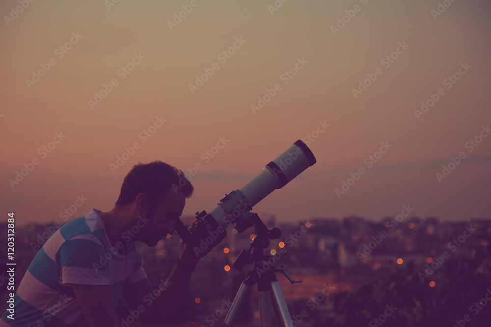 Guy looking at the stars with telescope and de-focused city lights in the background.