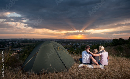 Father, mother and daughter near the tent sits with his back to the camera on a hill and hugging each other enjoying a beautiful sunset