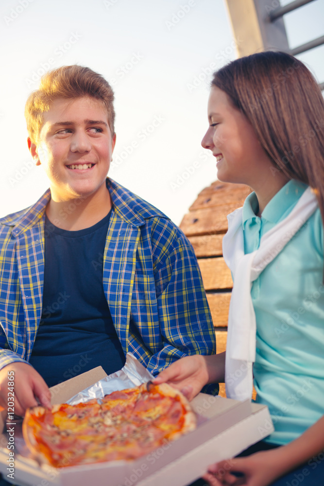 Two cheerful teenagers, girl and boy, eating pizza outdoor