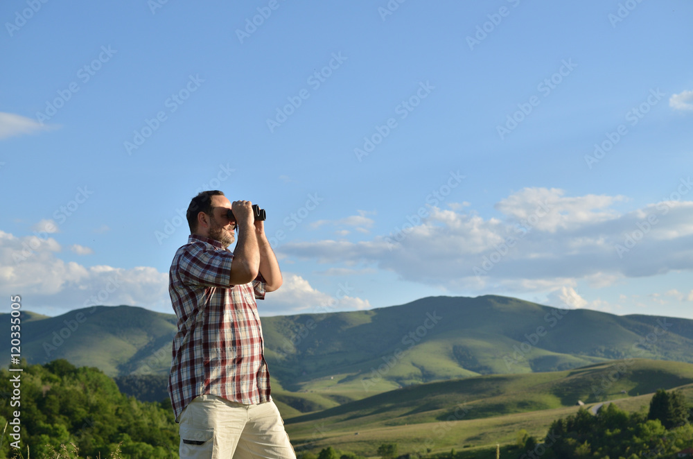 Man is using binoculars to watch divine summer scenery of green forest and grass on surrounding hills and mountain