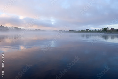 Morning nature scene: fog reflected in the water surface along with reeds, pink and blue sky, trees and a home on the shore. 