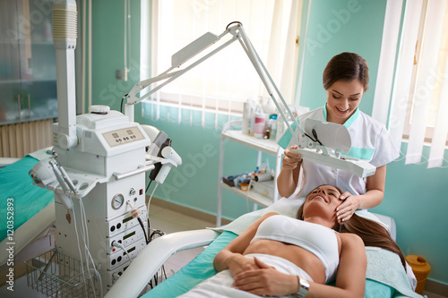 Fotografie Cheerful young girl is getting facial treatment by beautician