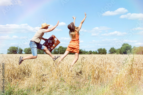 Joyful young couple having fun in wheat field. Excited man and woman running with retro leather suitcase on blue sky outdoor