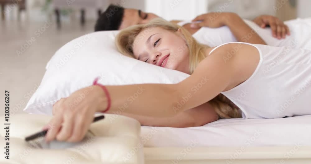 Woman turning off alarm clock cell phone lying in bed couple morning wake up smile Woman turning off alarm clock cell phone lying in bed couple morning wake up smile