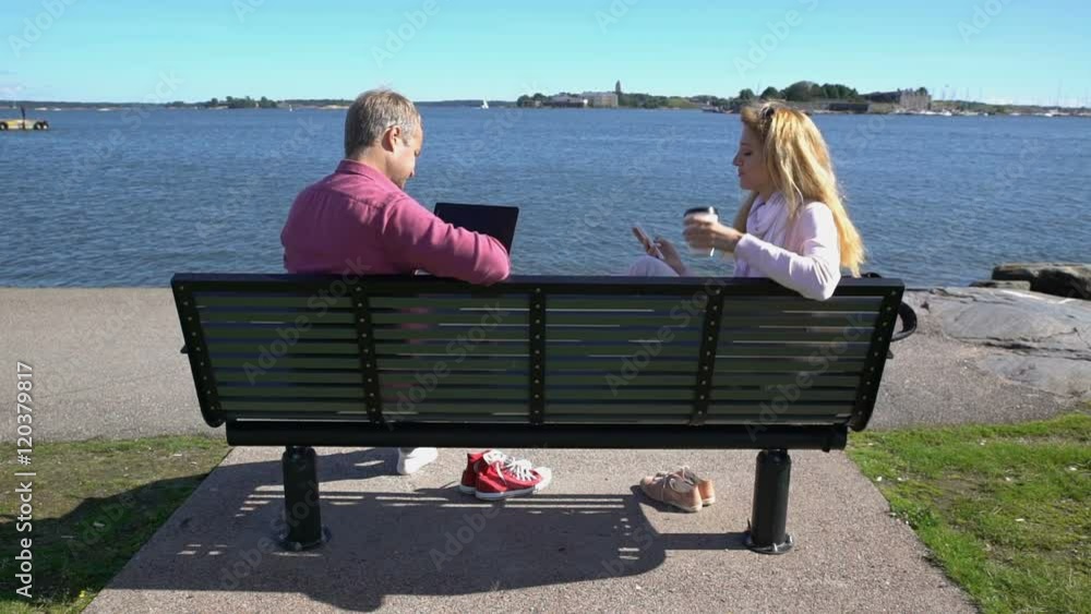 Couple on the bench by the sea. Young man using laptop, woman texting and drinking coffee. Slow motion.