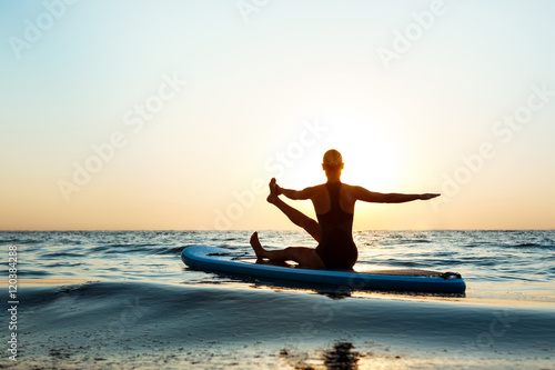 Silhouette of beautiful girl practicing yoga on surfboard at sunrise. © Cookie Studio