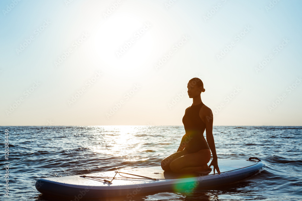 Silhouette of beautiful girl practicing yoga on surfboard at sunrise.