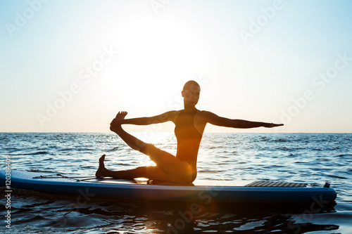 Silhouette of beautiful girl practicing yoga on surfboard at sunrise.
