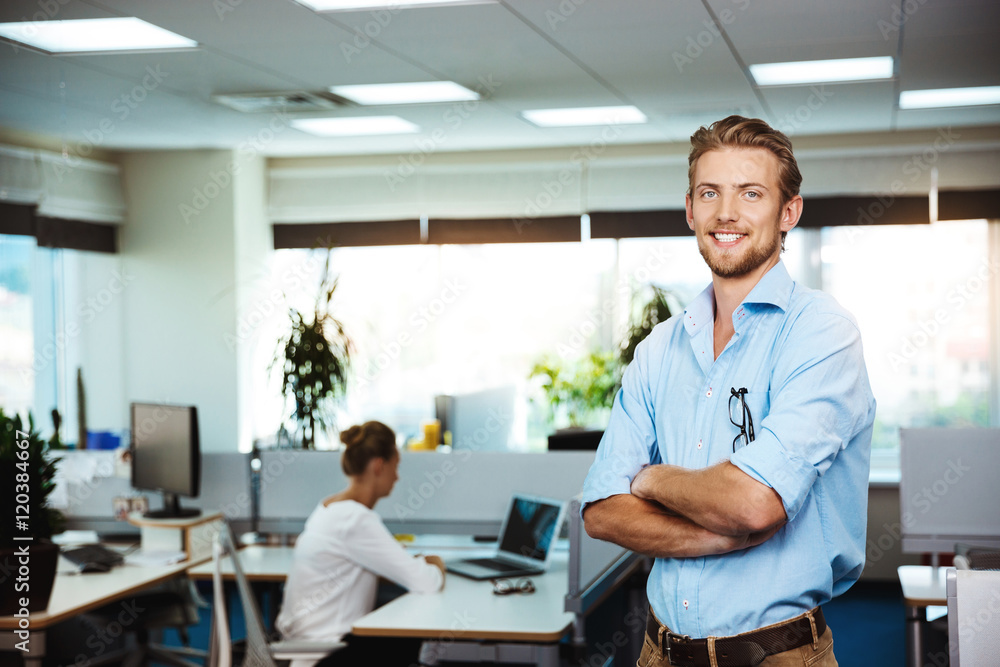 Young successful businessman smiling, posing with crossed arms, over office background.
