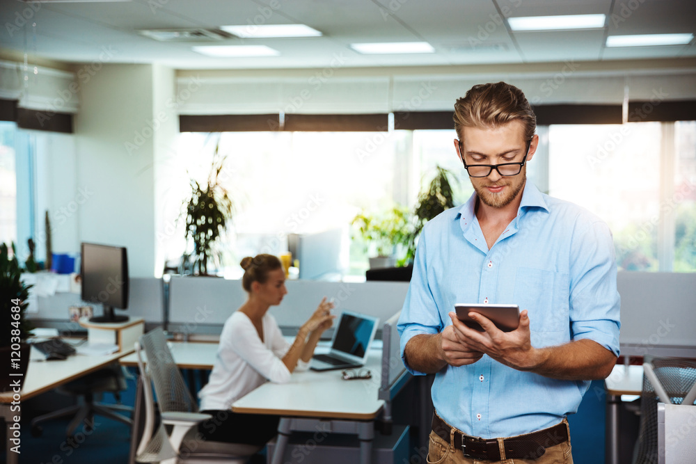 Young successful businessman smiling, looking at tablet, over office background. Young successful businessman smiling, looking at tablet, over office background.