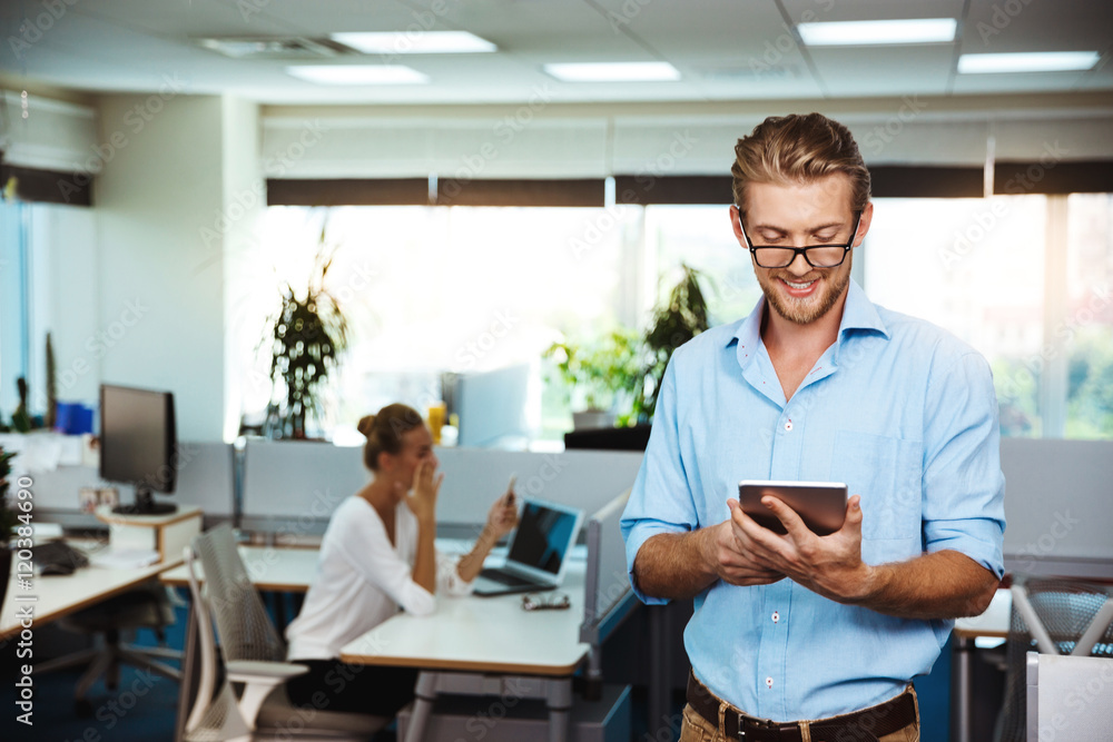 Young successful businessman smiling, looking at tablet, over office background. Young successful businessman smiling, looking at tablet, over office background.