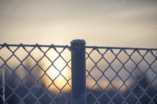A metal fence against a wintry sunset. Some snow and frost is covering the metal wires. 