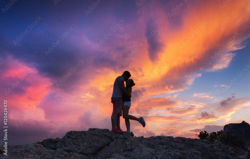 Landscape with silhouettes of a hugging and kissing man and girlfriend on the mountain peak at colorful sunset. Man and girl with beautiful sky. Silhouette of people. Couple, lovers, relationship.